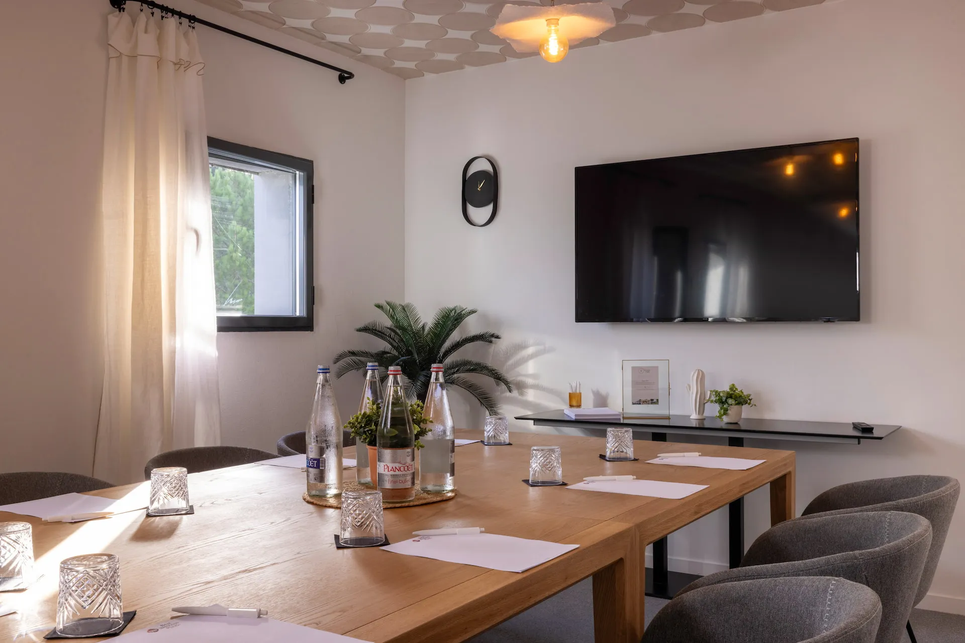 Modern meeting room with large wooden table, bottles of water, glasses, paper and pens, wall-mounted flat screen TV, black clock, and decorative plant near a window.