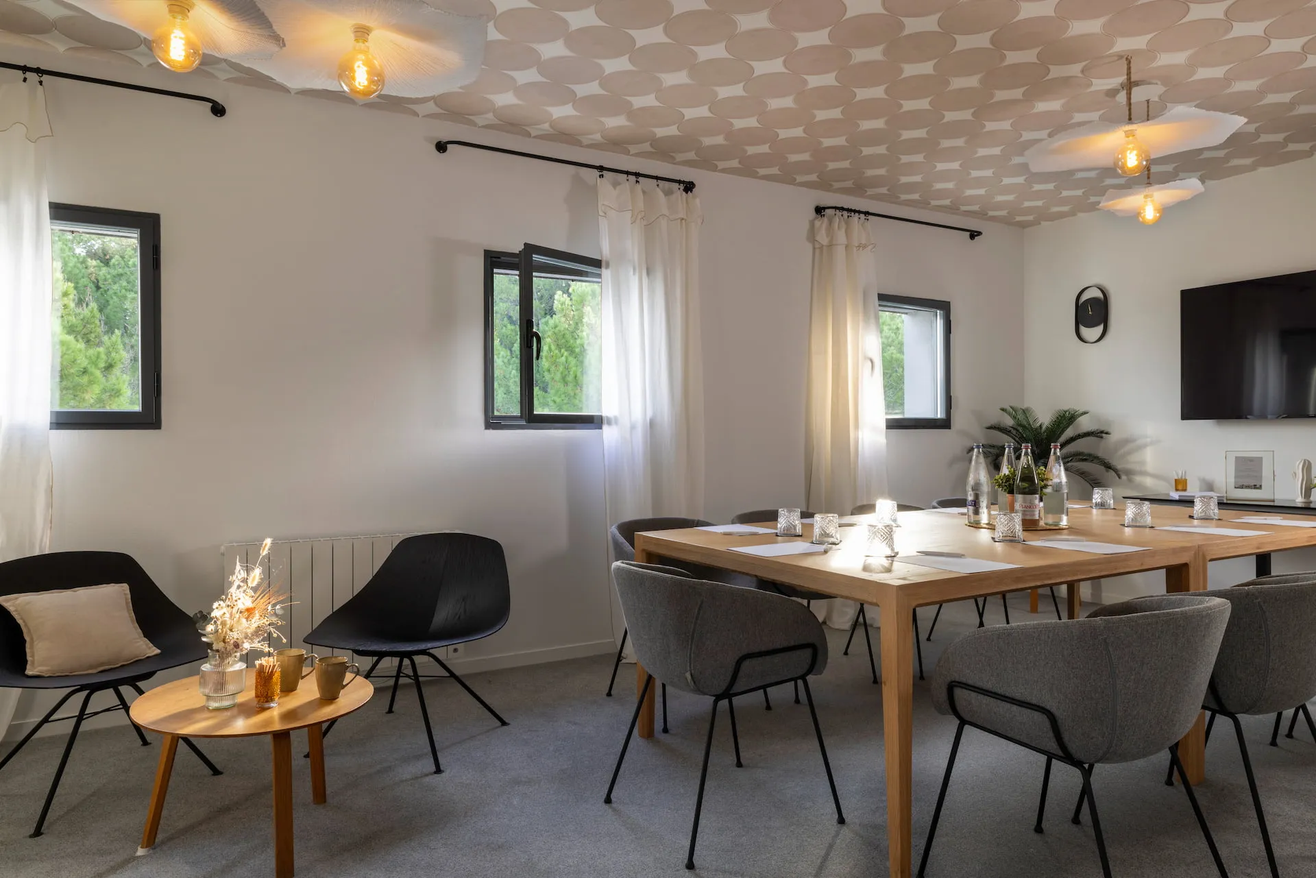 Bright meeting room with a large wooden table surrounded by gray chairs, two black armchairs with a small wooden table containing a floral decoration, bottles of water, and papers on the table.