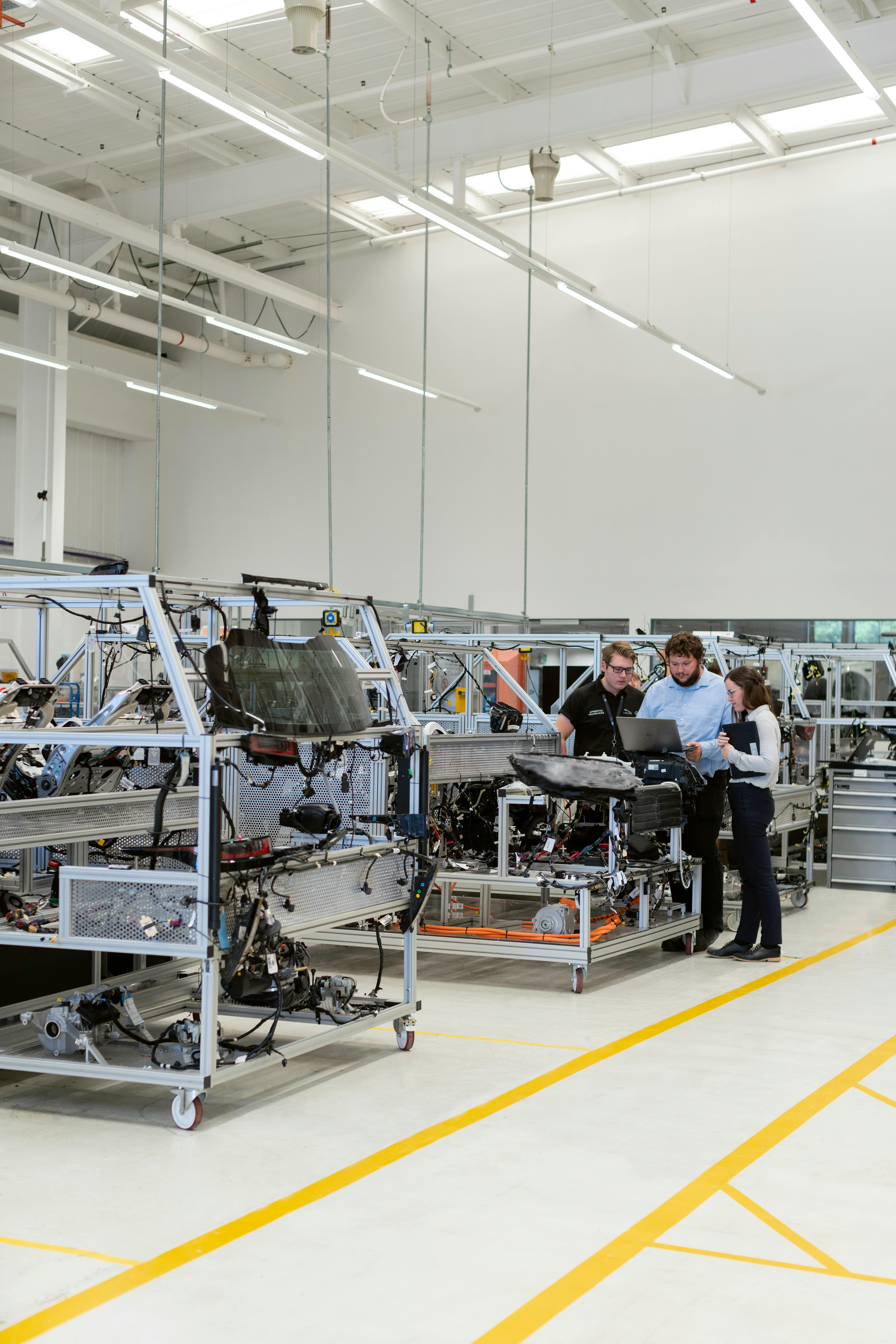 Three engineers collaborating and reviewing a laptop next to car chassis frames in a bright automotive workshop.