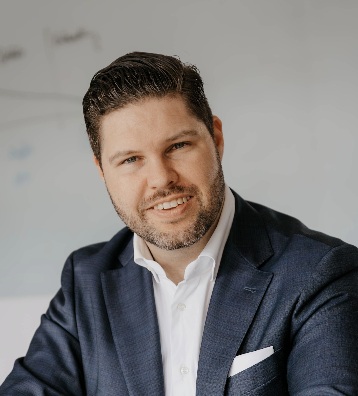 Smiling man with short dark hair and beard wearing a dark blue suit and white shirt.
