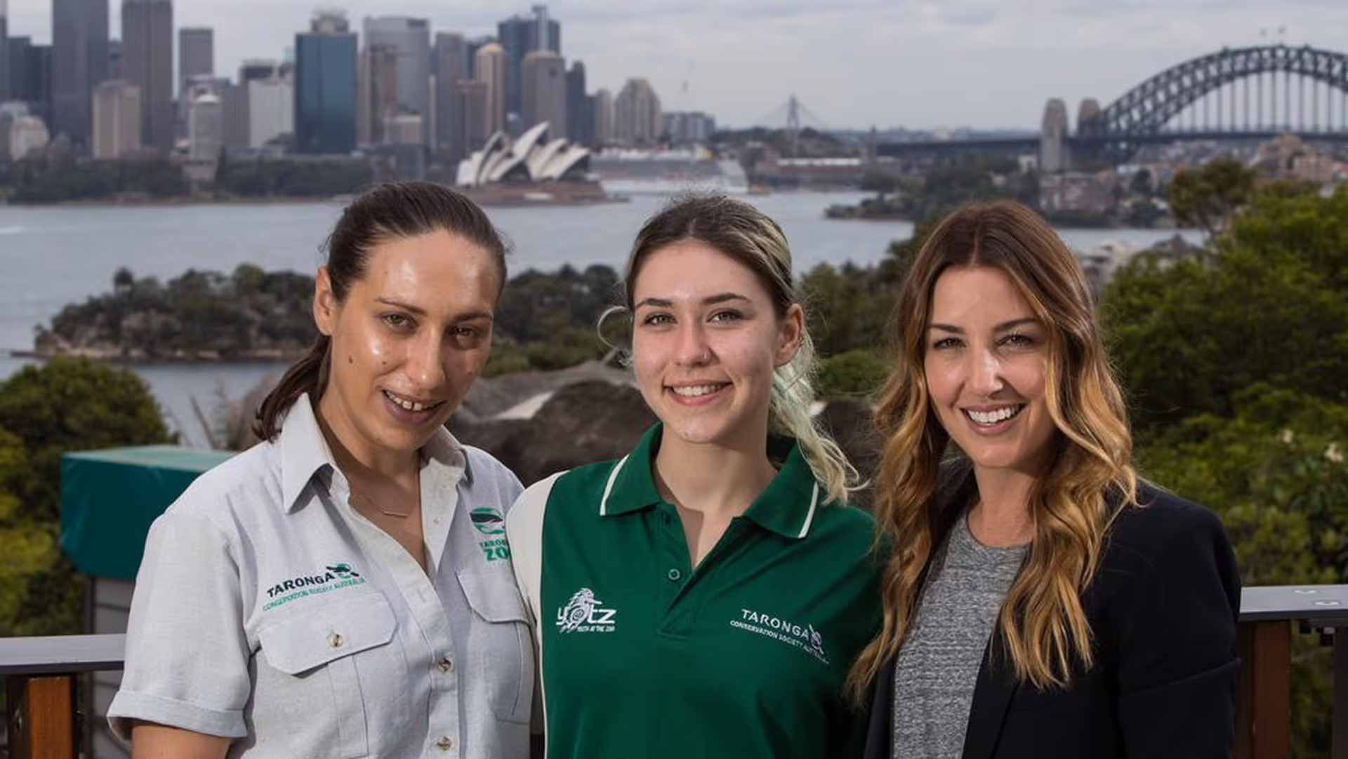 Three smiling women standing outdoors with Sydney Opera House and Sydney Harbour Bridge in the background.