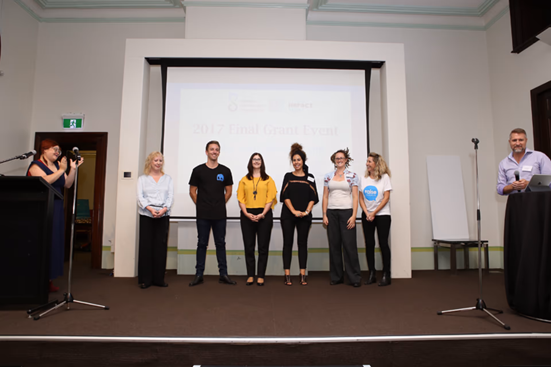 Seven people standing on stage in front of a presentation screen at the 2017 Final Grant Event, with one person clapping on the left and one person holding a microphone on the right.