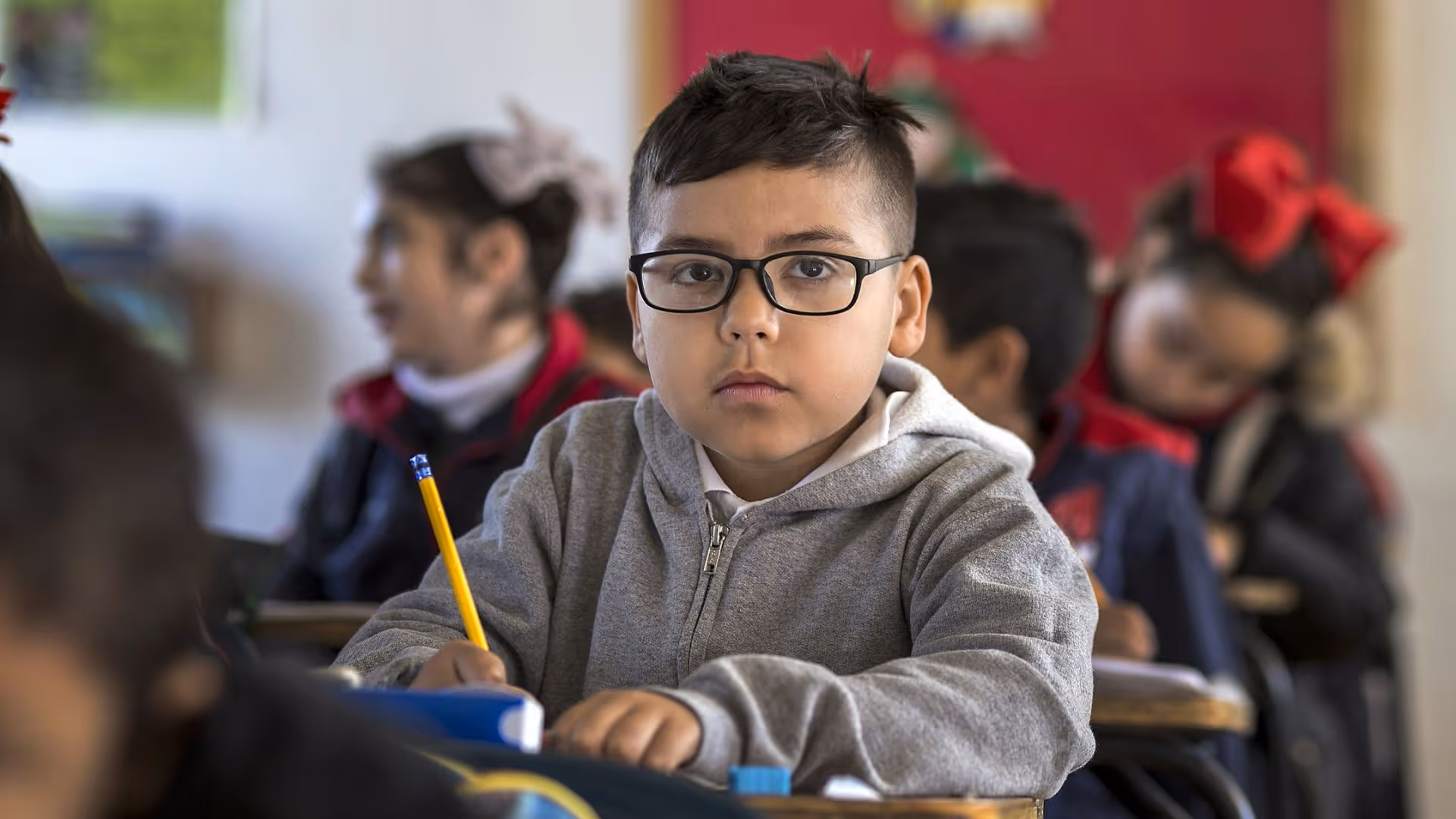 Young boy wearing glasses and a gray hoodie, sitting at a desk in a classroom holding a pencil.