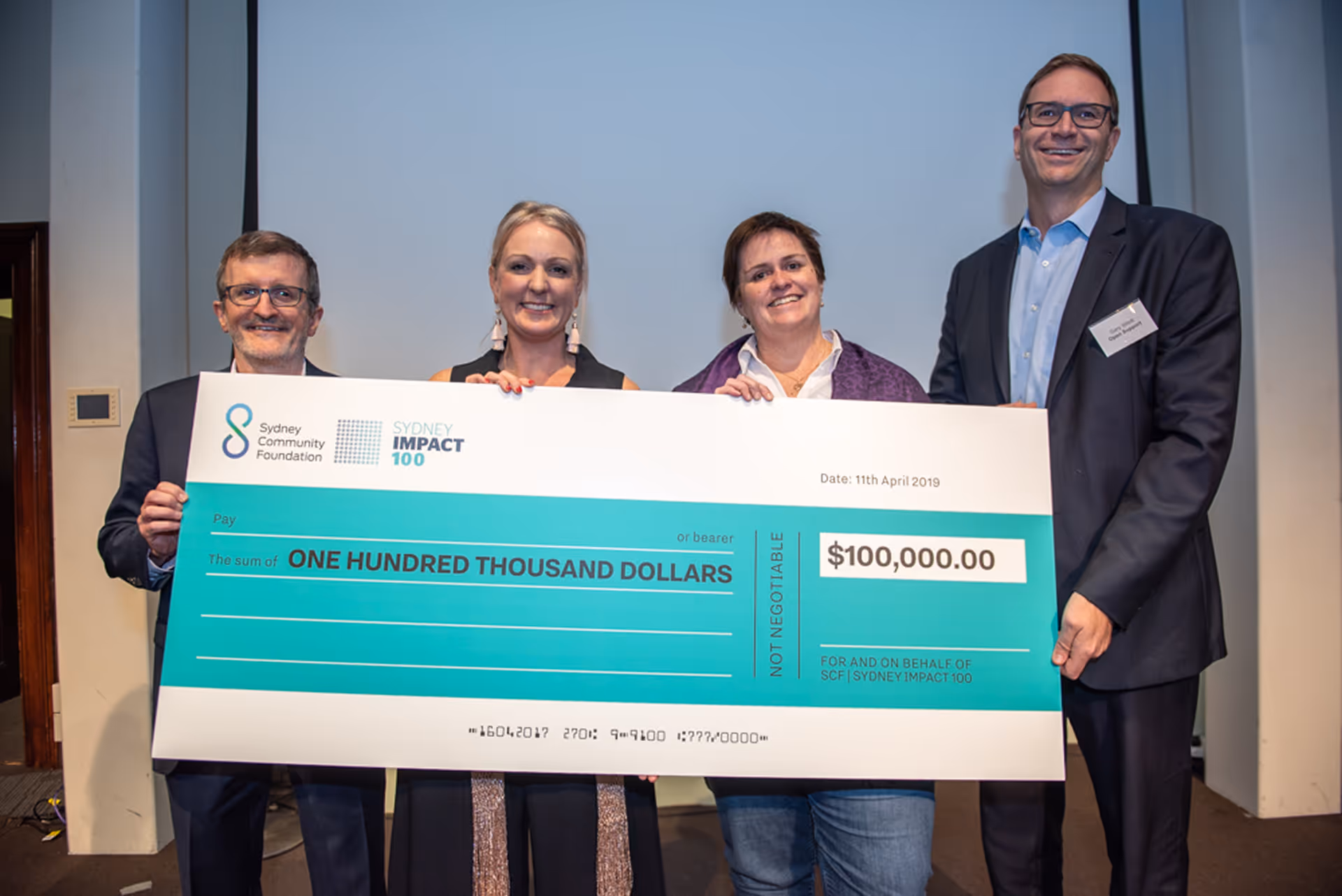 Four people holding a large ceremonial check for $100,000 from Sydney Community Foundation and Sydney Impact 100, dated 11th April 2019.