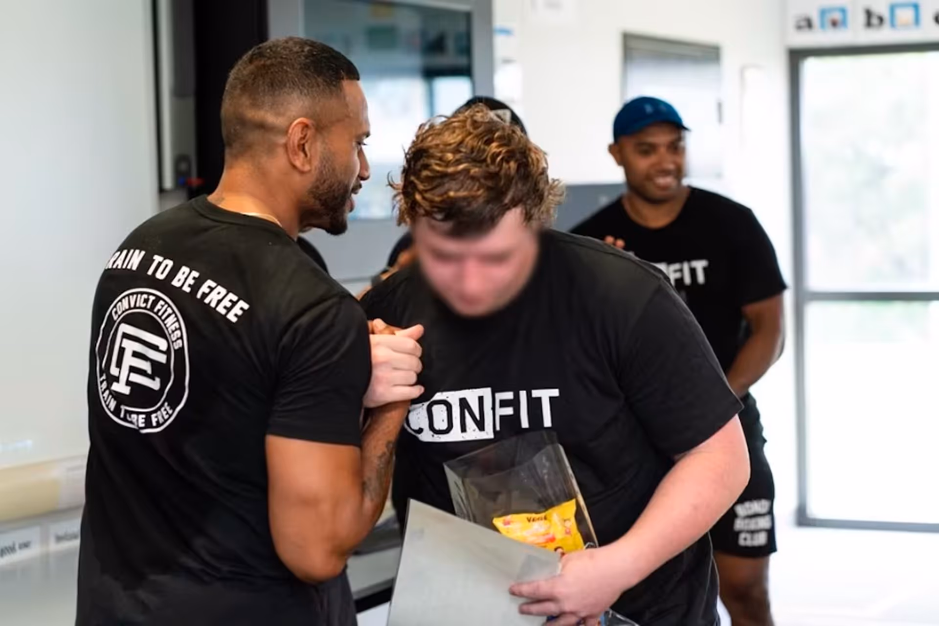 Two men in black t-shirts shaking hands, one holding an award, with another man smiling in the background inside a bright room.