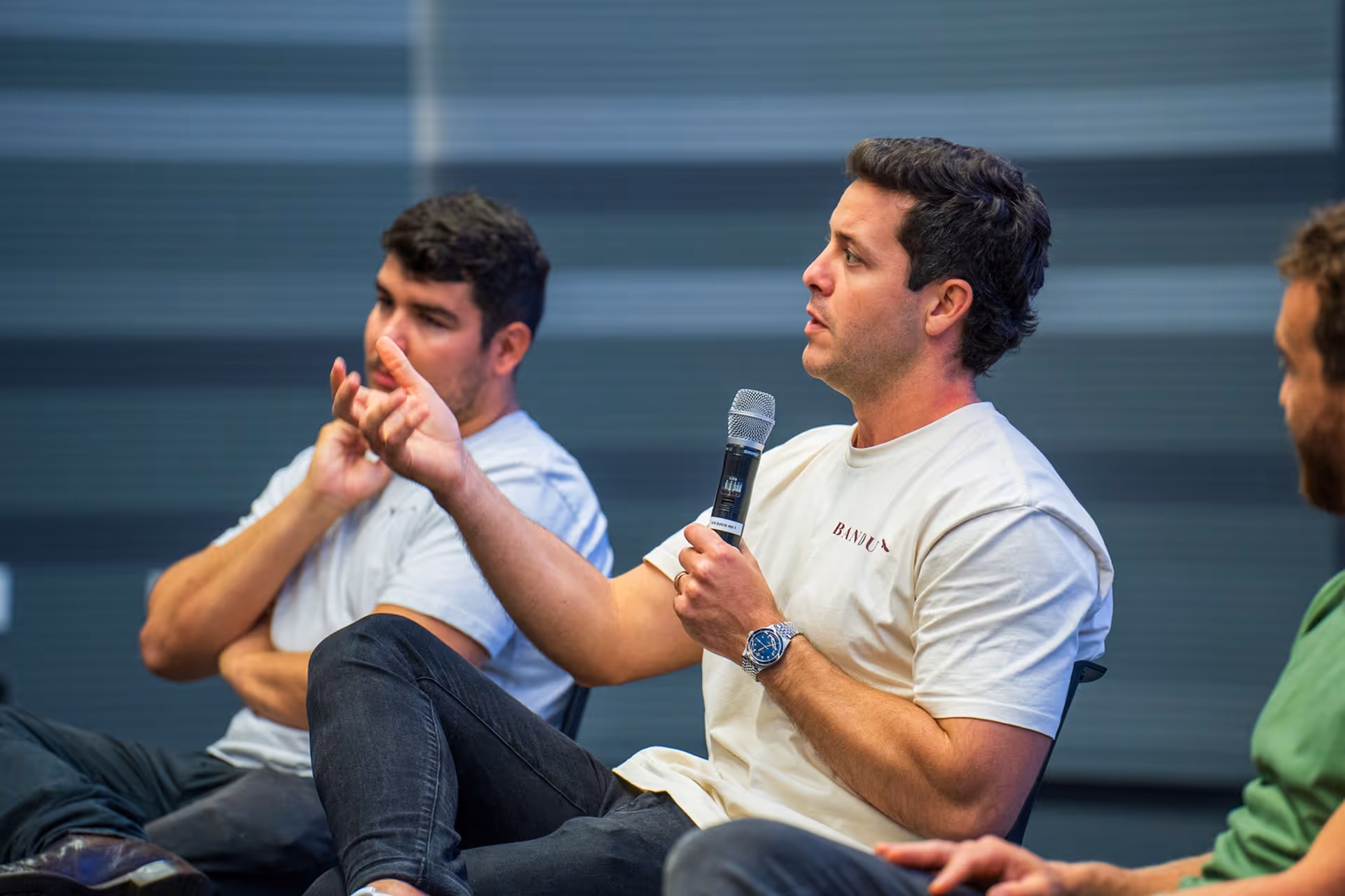 Three men engaged in a discussion, one speaking into a microphone while gesturing with his hand.