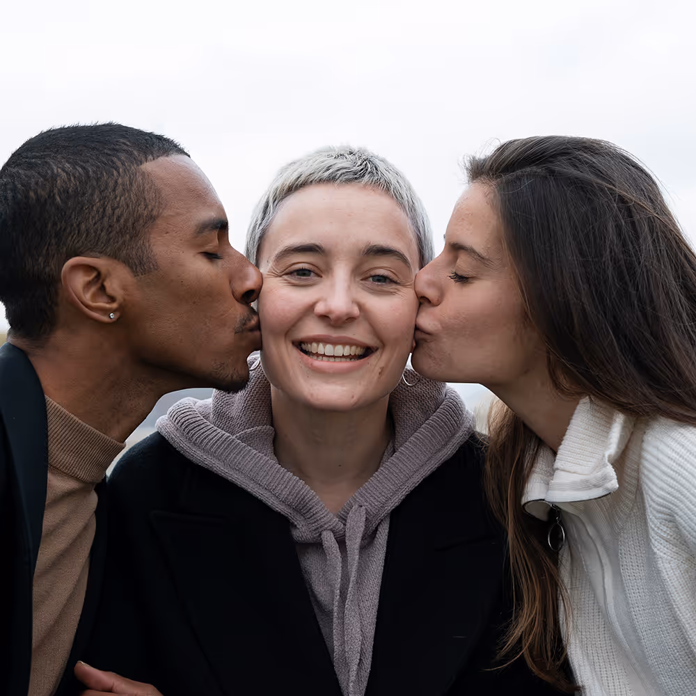 Smiling person with short blonde hair being kissed on each cheek by two friends outdoors.