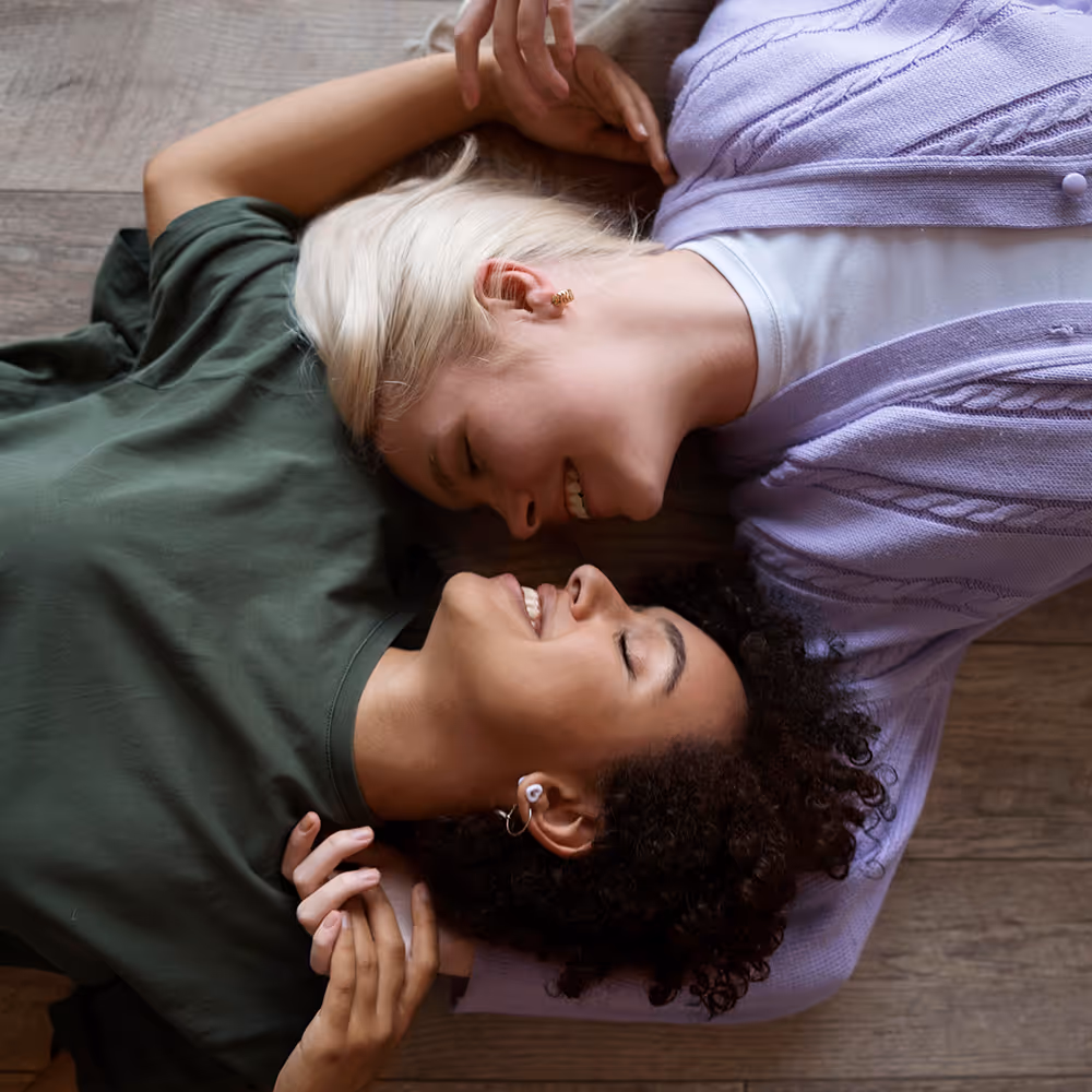 Two women lying on the floor facing each other, smiling and holding hands.