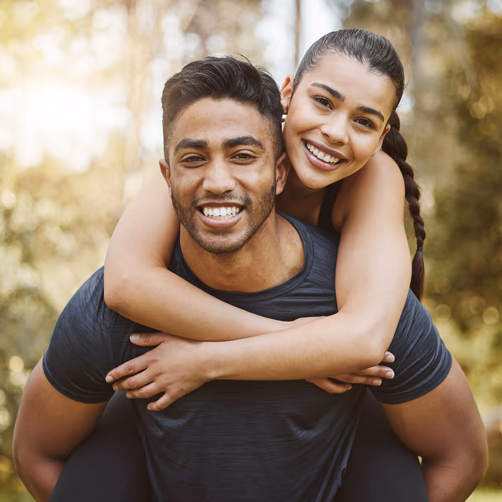 Smiling young woman with braided hair giving a piggyback ride to a smiling young man outdoors in a sunlit park.