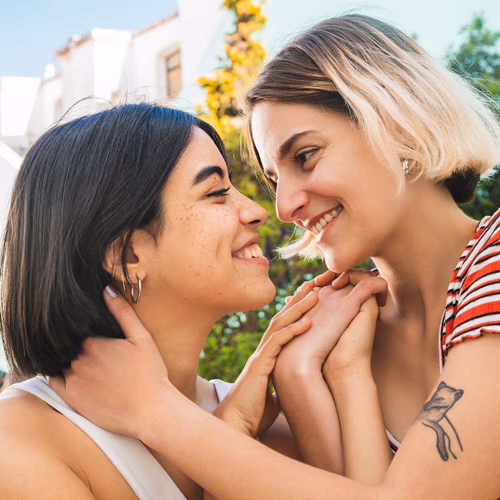 Two women smiling closely and holding hands affectionately outdoors.