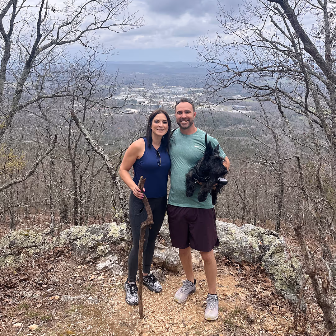 Smiling couple standing on a hiking trail clearing with leafless trees, holding a walking stick and a small black dog.