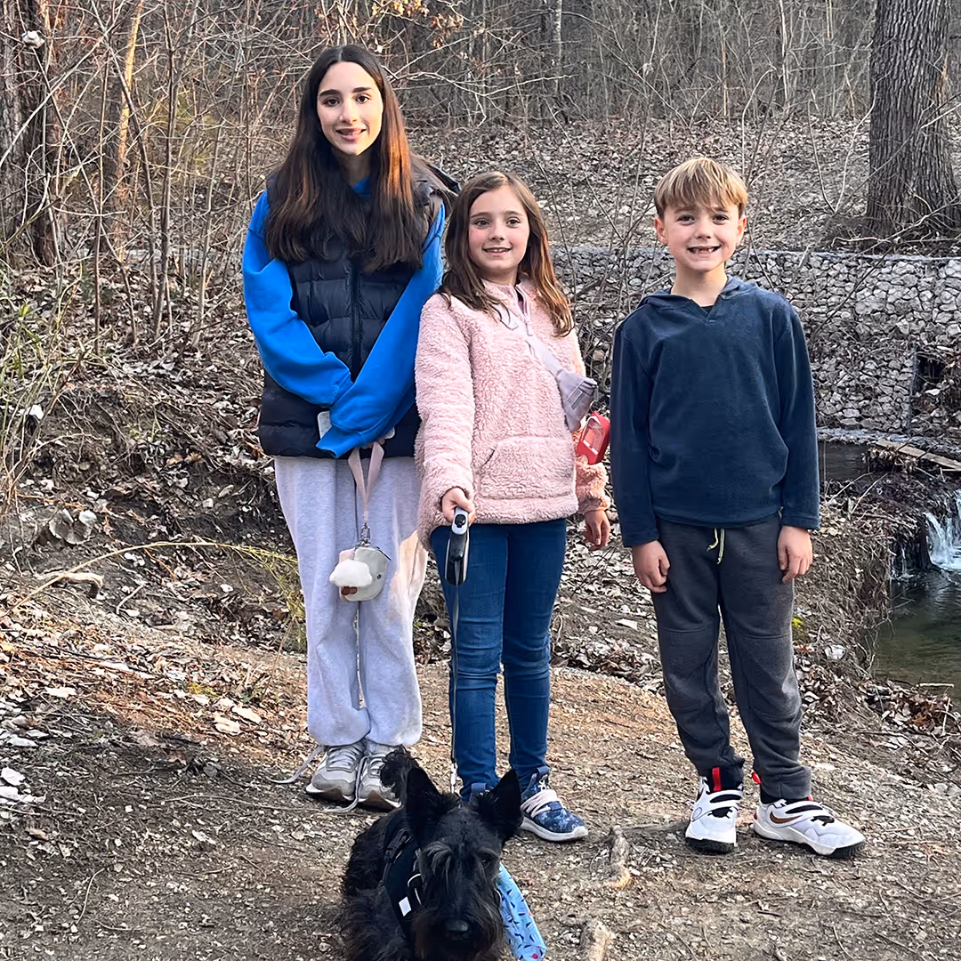 Three children standing outdoors in a wooded area with a black small dog sitting in front of them.