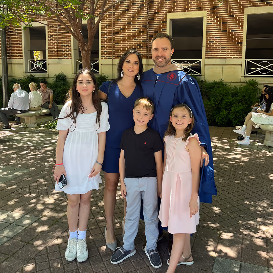 A family of five posing outdoors on a paved area with a brick building in the background, including a man in graduation gown, a woman in a navy dress, two girls in white and pink dresses, and a boy in a black shirt and gray pants.