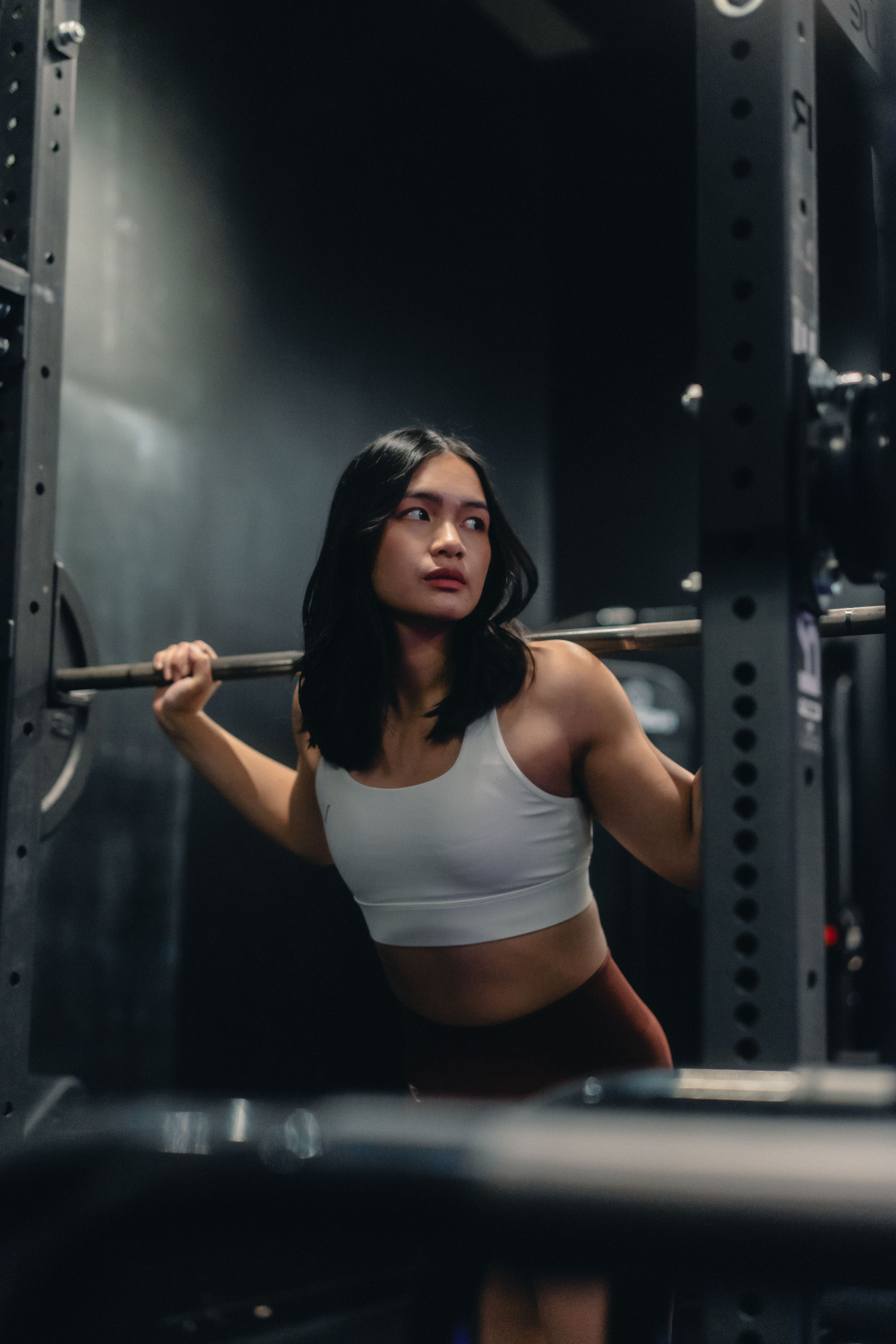 Woman in a white sports bra preparing to lift a barbell in a gym squat rack.