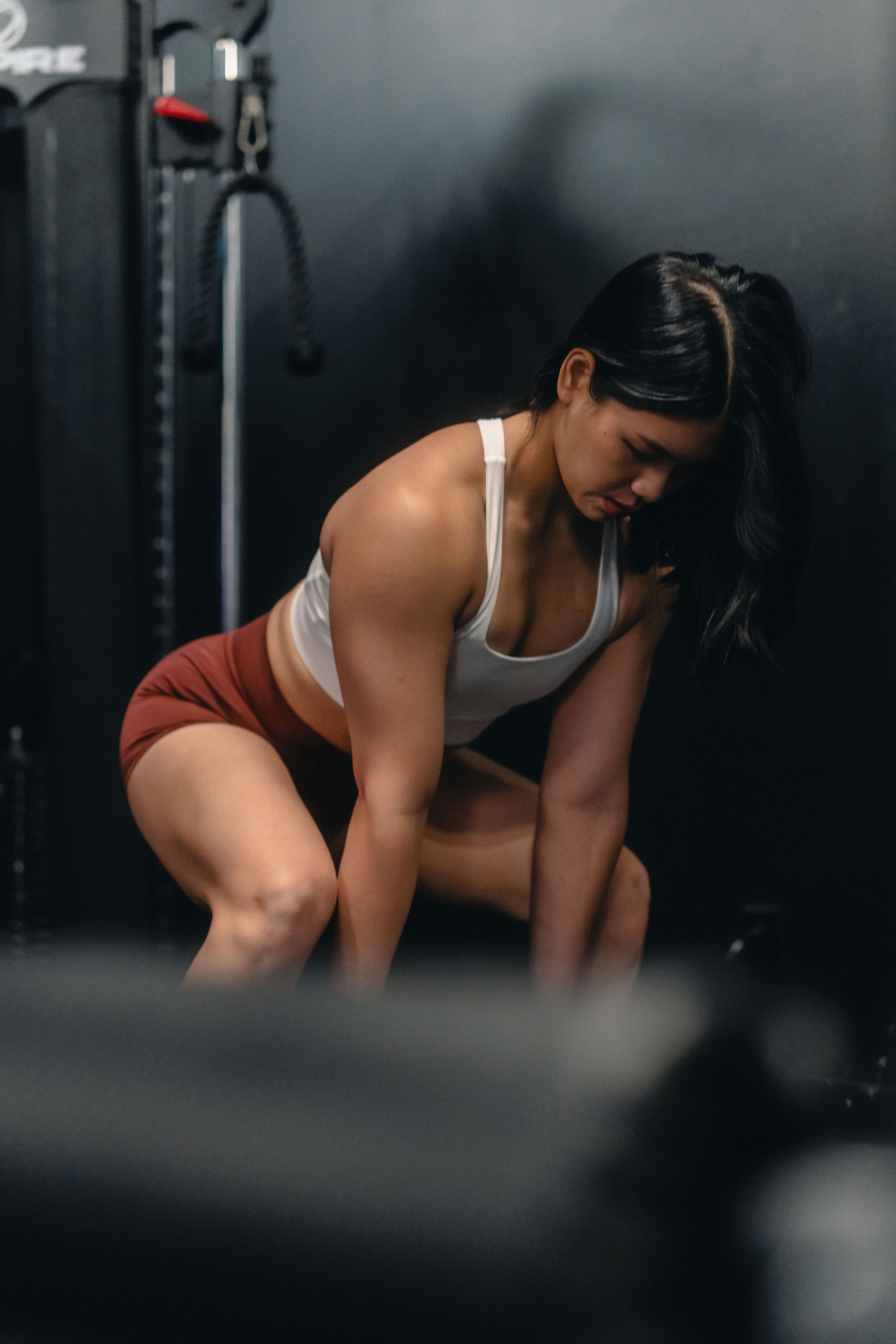 Woman in white sports bra and red shorts bending forward during a workout in a gym with dark walls.