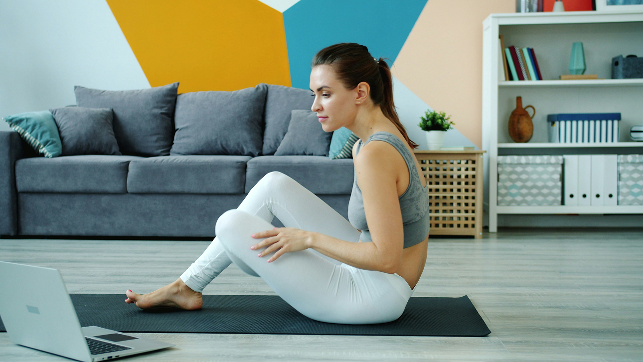 Woman in workout clothes sitting on a yoga mat in a living room following an exercise on a laptop.