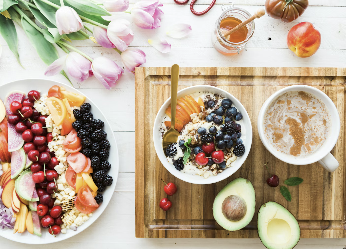 Breakfast spread with a bowl of yogurt topped with blueberries, blackberries, cherries, nuts, and peach slices, a cup of coffee with cinnamon, half an avocado, a plate of assorted fresh fruits and vegetables, and pink tulips on a white wooden table.