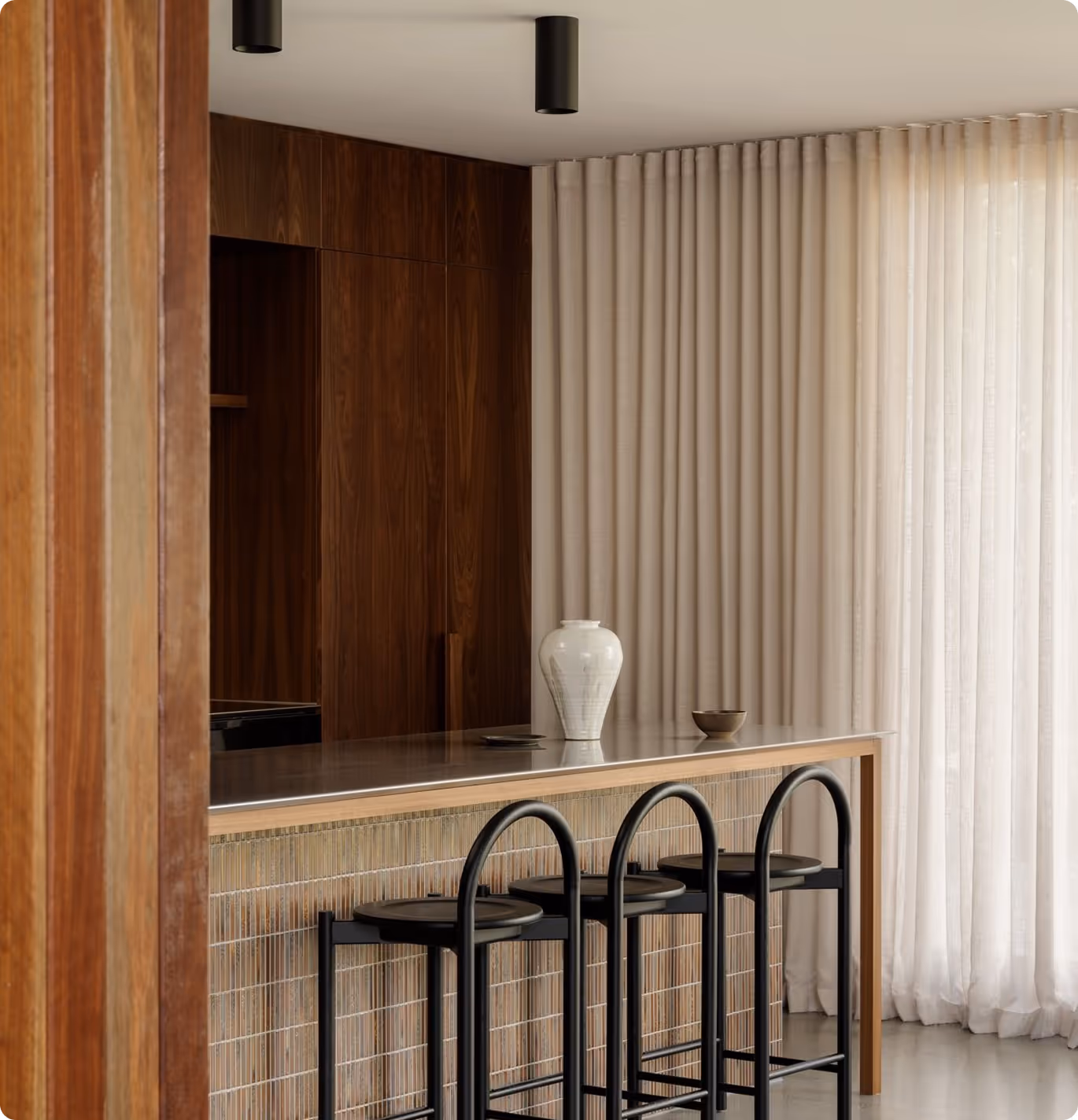 Modern kitchen island with three black bar stools, a white vase, and a small bowl in front of beige curtains and dark wood cabinets.