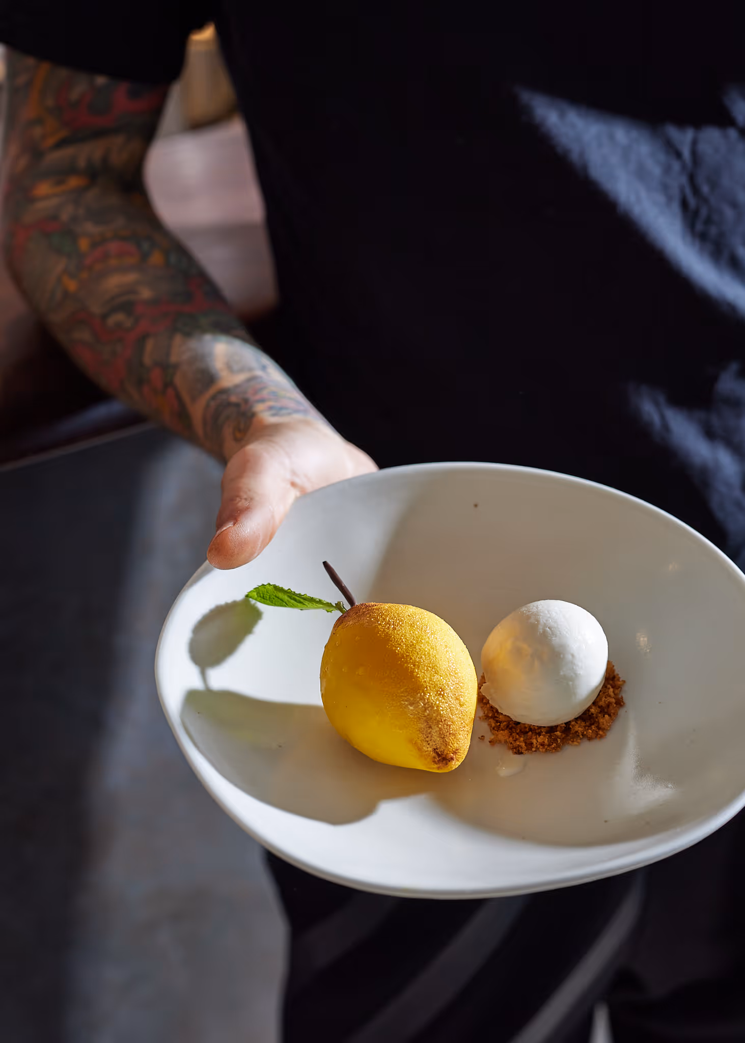 Tattooed arm holding a white plate with a yellow dessert shaped like a pear with a leaf, and a white ice cream scoop on crumbs.