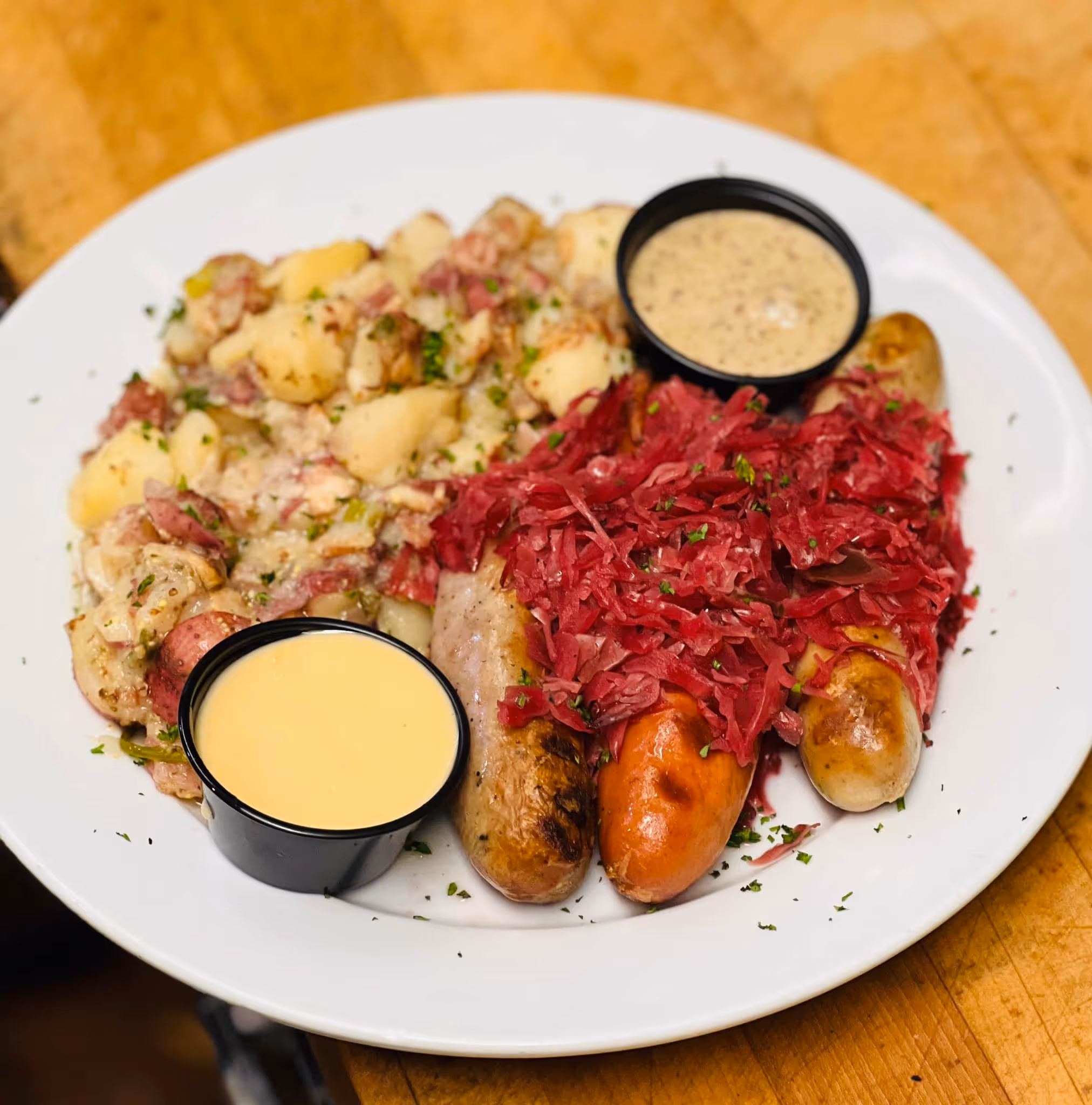 A plate of food with a black bowl of sauce and a white plate with a sausage and potatoes.
