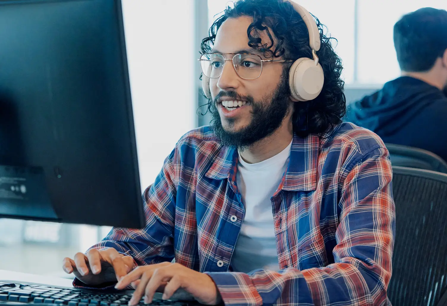 Smiling man wearing glasses and headphones typing on a keyboard while looking at a monitor in an office.