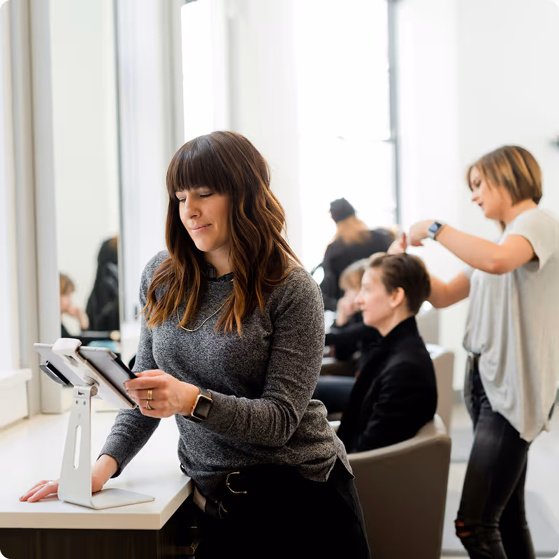 Woman with long brown hair using a tablet at a salon reception while another woman styles a customer's hair in the background.