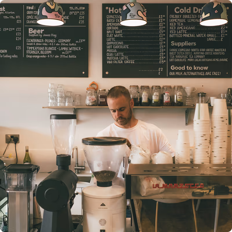 Barista preparing coffee behind a counter with coffee grinders, stacked cups, and chalkboard menu displaying drinks and prices.
