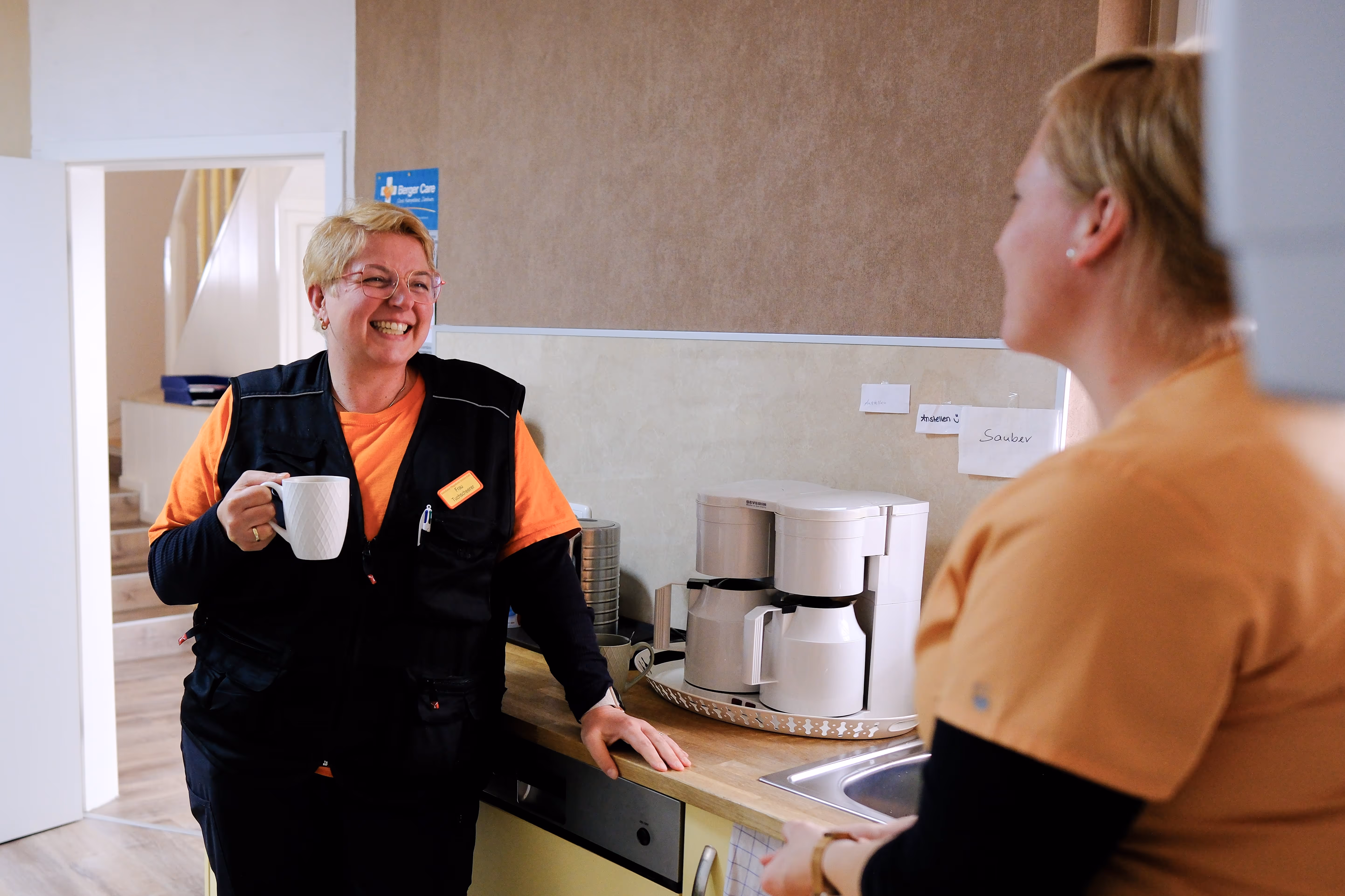 Pflegekraft in orange-schwarzer Arbeitskleidung trinkt Kaffee und spricht lächelnd mit Kollegin in der Teeküche