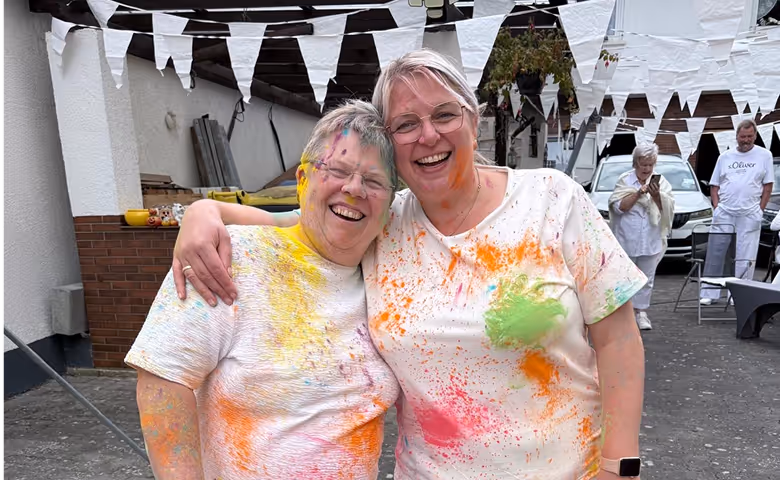 Zwei lachende Frauen umarmen sich beim Firmenfest – bunte Farbpulver-Action unter Wimpelketten im Hof