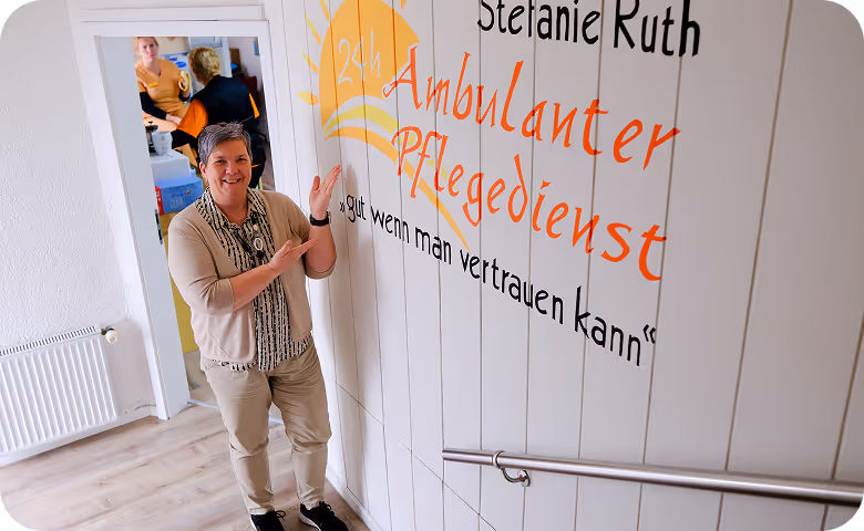 Frau steht im Treppenhaus neben dem Logo „Stefanie Ruth – Ambulanter Pflegedienst“ und zeigt darauf