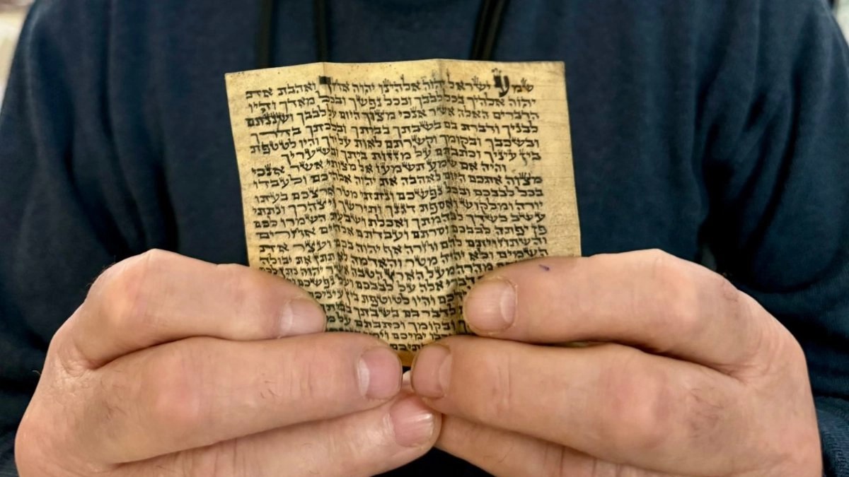 A pair of hands holds open a small, aged parchment mezuzah scroll inscribed with Hebrew text from the Shema prayer, its yellowed surface and hand-lettered script showing decades of wear from being carried in a wallet during the Soviet era.