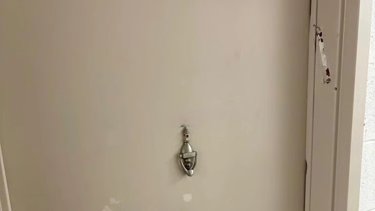 A beige door with a small silver door knocker, showing faint scuff marks on its surface and traces of adhesive residue on the right-side doorframe where a mezuzah was once been affixed.