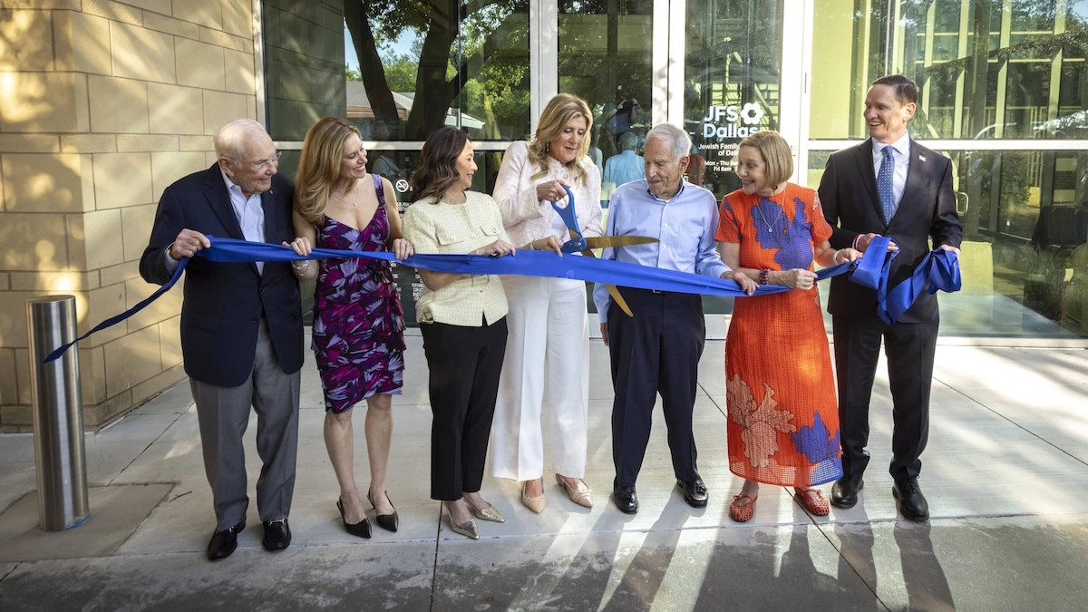 Community leaders and donors cut a blue ribbon with gold scissors at the grand opening of the new JFS Dallas Steinberg Building, standing in front of the glass entrance with the Jewish Family Service of Dallas sign visible behind them
