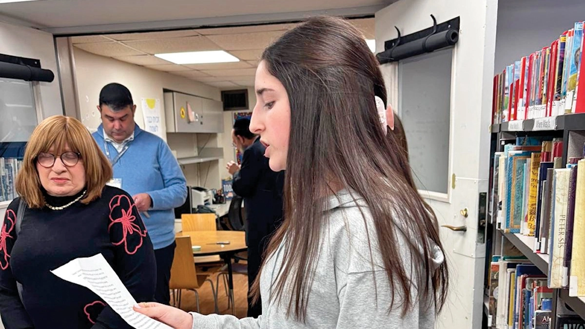 A student reads aloud from a printed sheet during a mezuzah dedication ceremony at Moriah School in Englewood, with Morah Chaya Devora and other staff members looking on in the school library surrounded by bookshelves