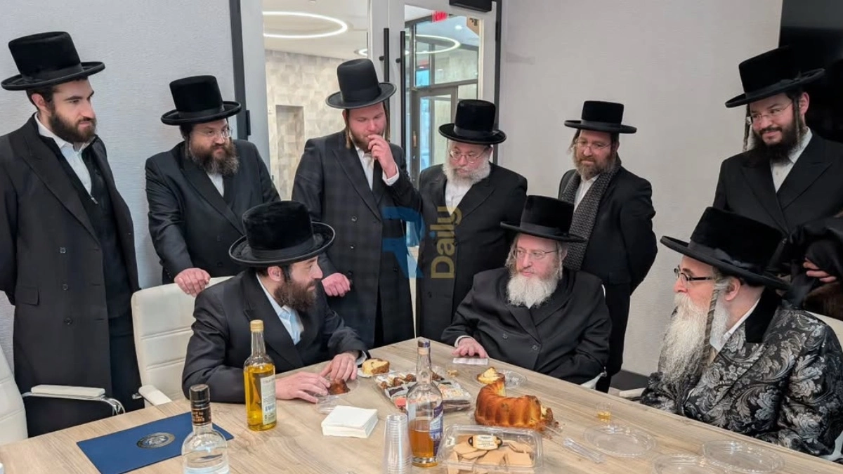 Hasidic rabbinic leaders and community members in black hats and bekeshes gather around a conference table with challah, schnapps, and refreshments at the new Yedei Chesed headquarters in Rockland County following a kevias mezuzah ceremony, with the Rockla