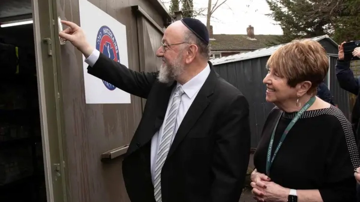 Chief Rabbi Ephraim Mirvis reaching up to affix a mezuzah on a doorpost, with a smiling woman beside him at the Hatzola HBS cabin in Barnet