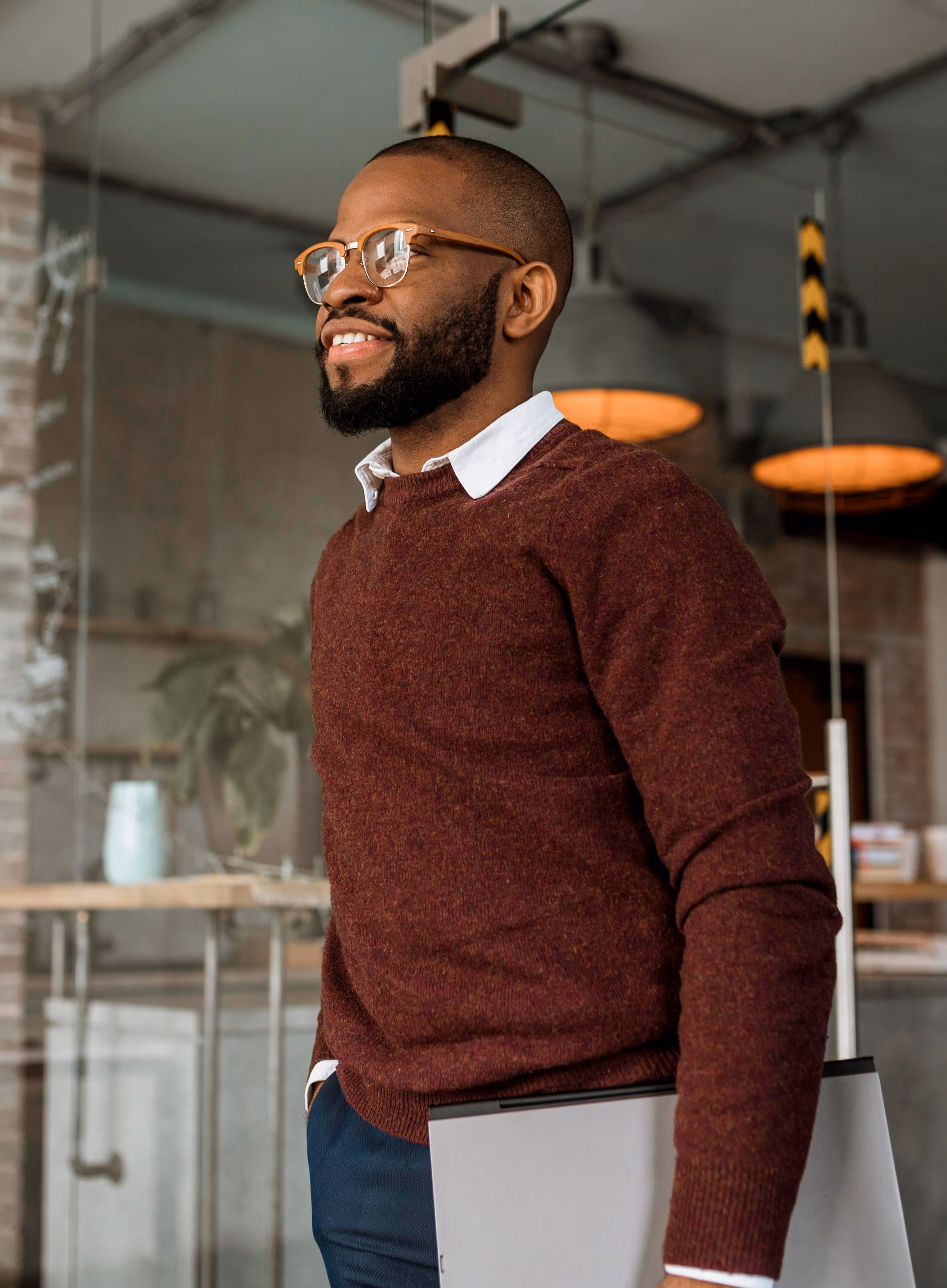 Smiling man wearing glasses and a maroon sweater holding a laptop in a modern office.