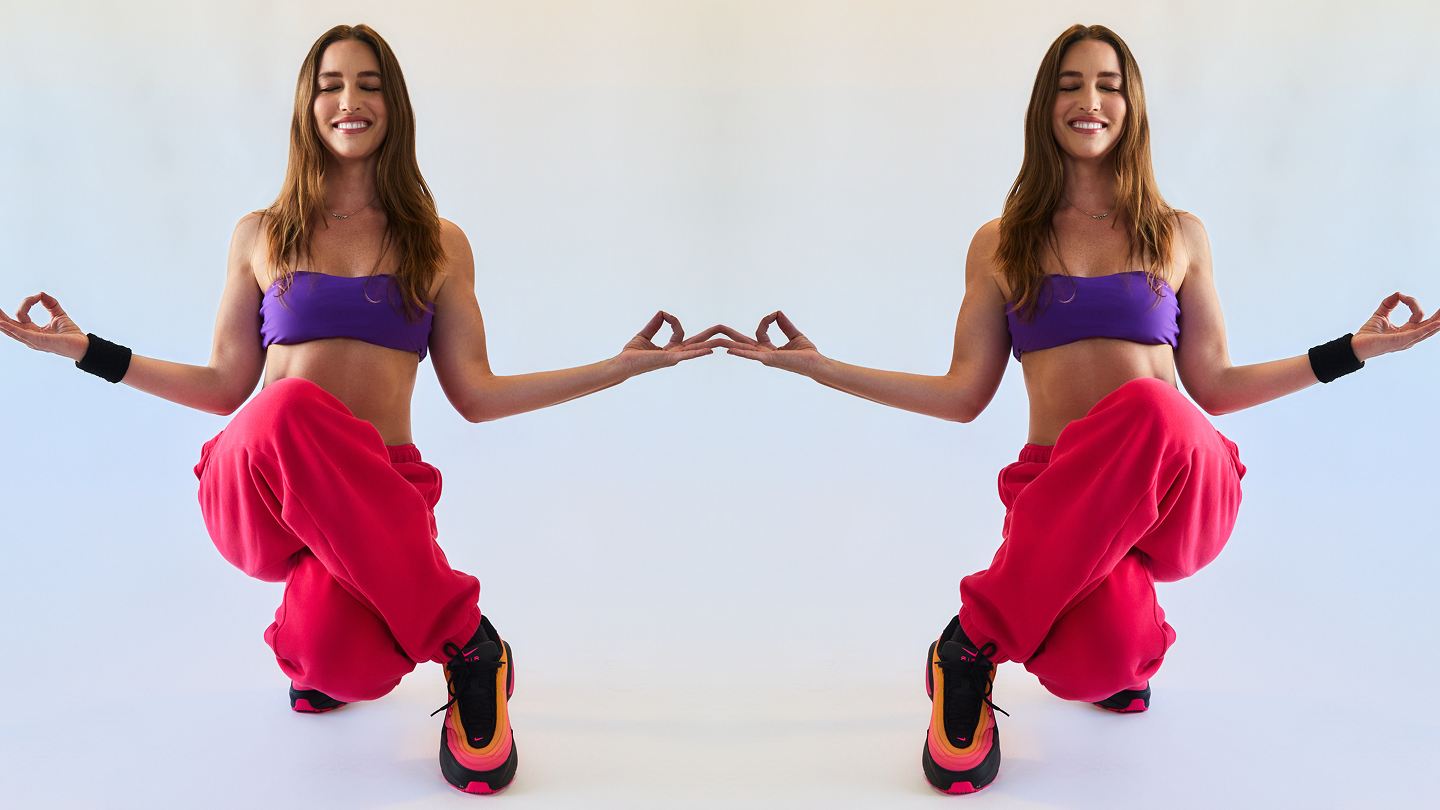 Mirrored image of a smiling woman meditating in a cross-legged squat, wearing a purple crop top, red pants, and colorful sneakers.