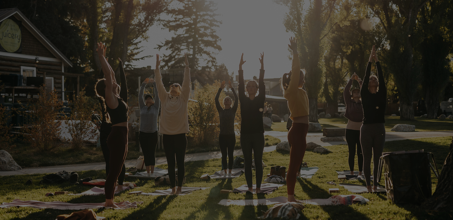 Group of women practicing yoga outdoors on mats with arms raised towards the sky in a park setting during sunset.
