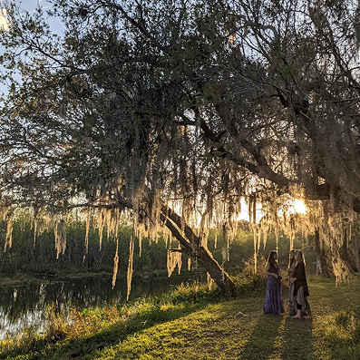 Two women standing and talking under a large tree with hanging moss near a calm river at sunset.