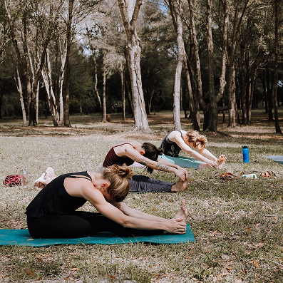 Three women seated on yoga mats outdoors in a park stretching forward to touch their toes.