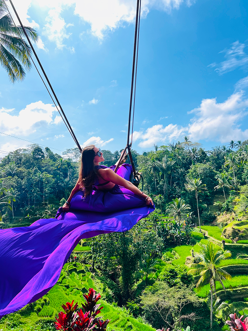 Woman in a flowing purple dress swinging over a lush green terraced landscape under a bright blue sky with scattered clouds.