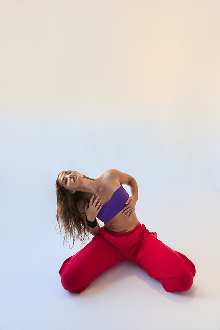 Woman with long hair kneeling and leaning back, wearing a purple tube top and red pants, against a light gradient background.