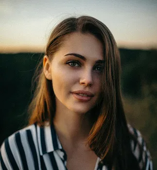Portrait of a young woman with long brown hair wearing a black and white striped shirt, with a blurred nature background.