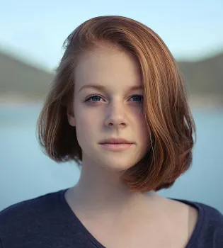 Portrait of a young woman with short reddish-brown hair and blue eyes against a blurred outdoor background.
