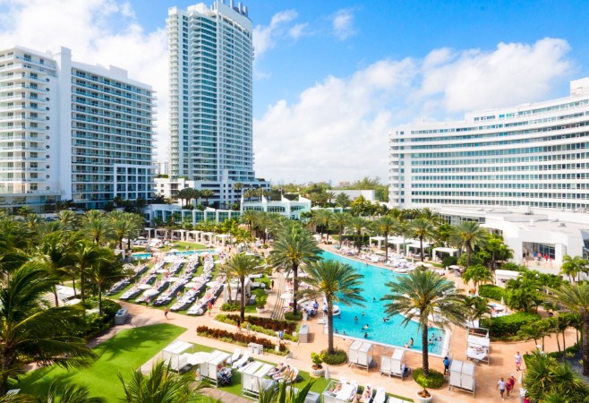 A sunny resort with tall white hotel buildings surrounding a large swimming pool and numerous lounge chairs, amidst palm trees and greenery.