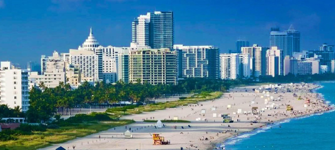 Aerial view of a wide sandy beach with scattered lifeguard towers and people, bordered by palm trees and tall modern buildings under a clear blue sky.