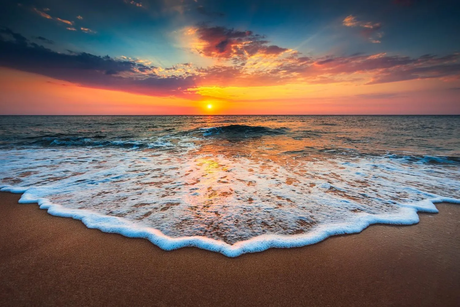 Sunset over ocean waves gently washing onto sandy beach under a colorful sky.