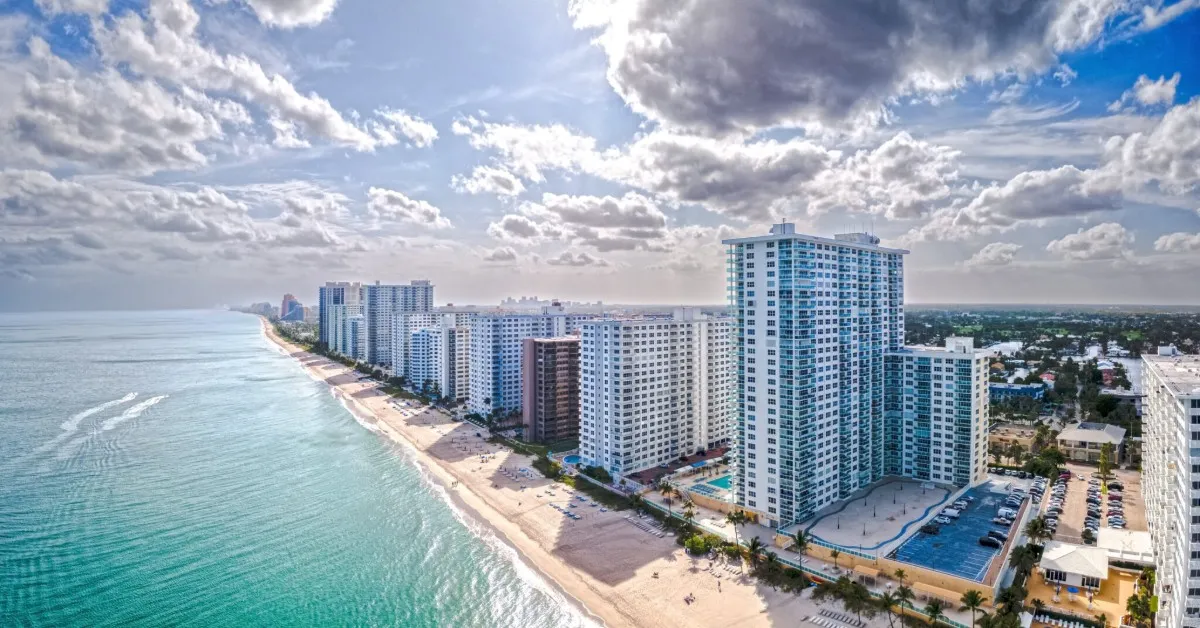 Aerial view of tall beachfront condominium buildings alongside a sandy beach with turquoise ocean water under a partly cloudy sky.