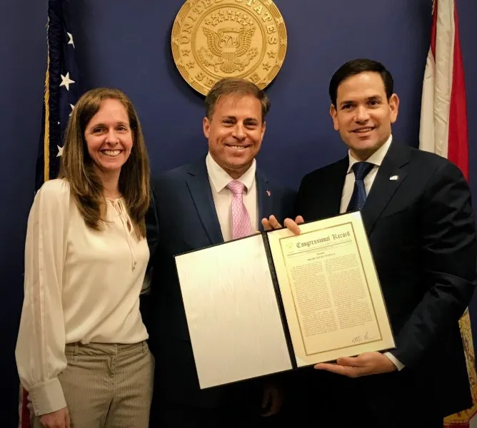 Three individuals smiling, with the man on the right holding an open framed document titled 'Congressional Record' in front of a U.S. government seal and flags.